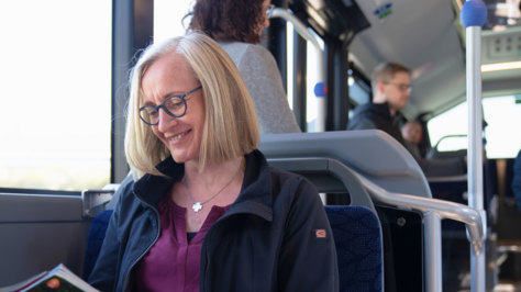 Lächelnde Frau sitzt in einem Linienbus und liest eine Zeitschrift.